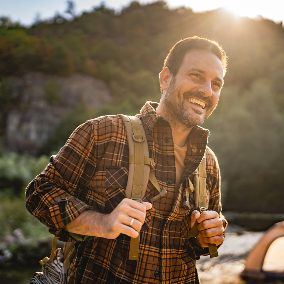 Man wearing a plaid shirt and backpack smiles outdoors in a sunlit, wooded area with a tent in the background.