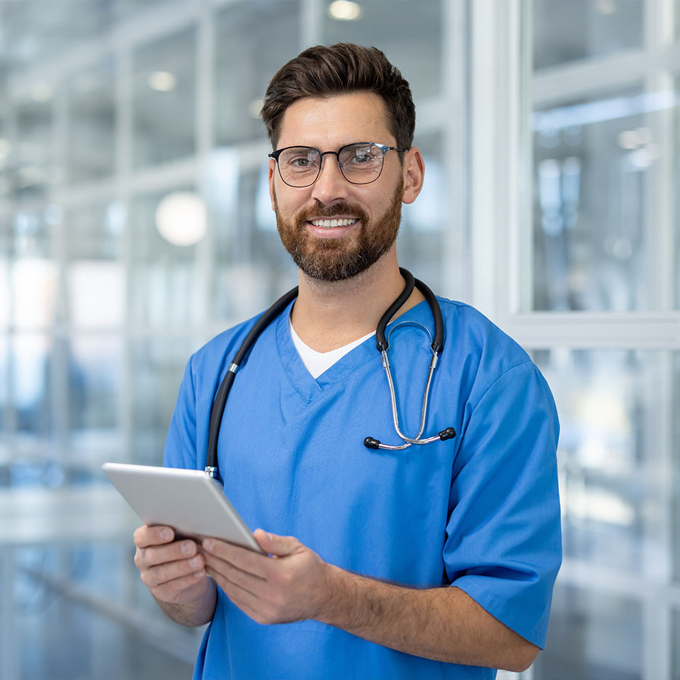 A male healthcare professional in blue scrubs and glasses holds a tablet while standing in a modern medical facility.
