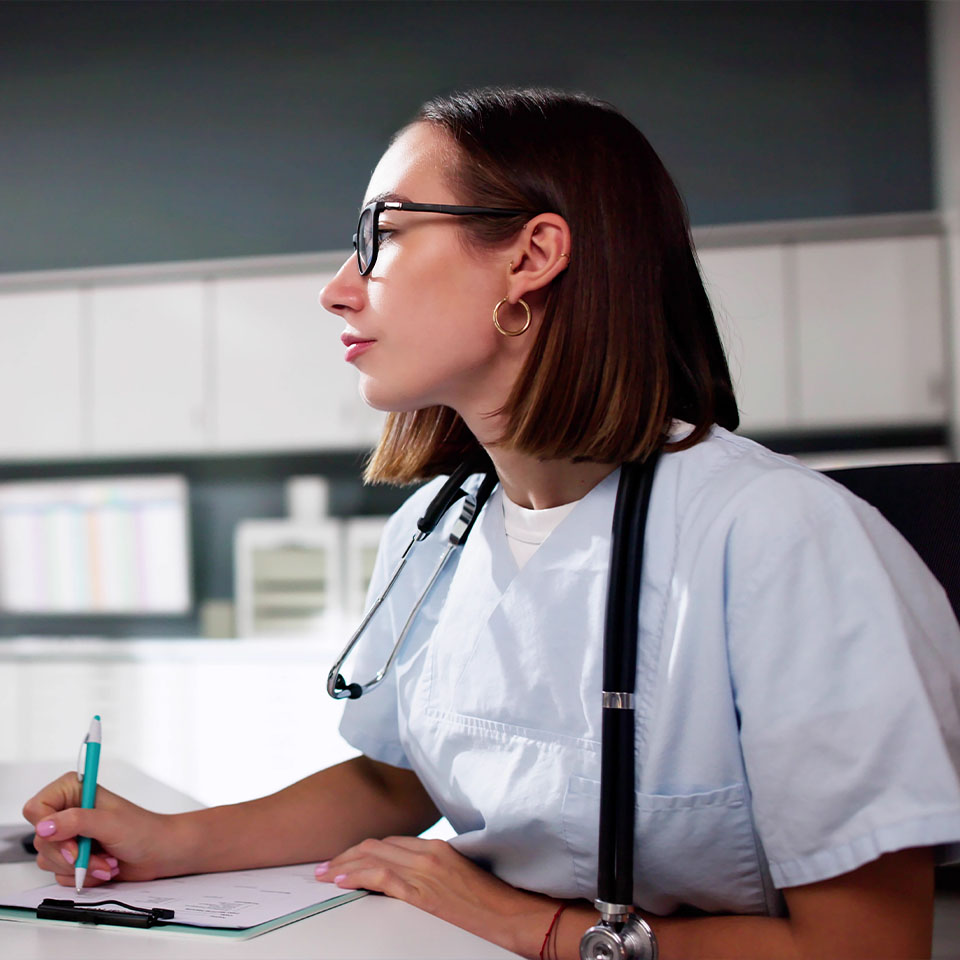 A healthcare professional wearing scrubs and a stethoscope writes notes on a clipboard while seated at a desk in a medical office.