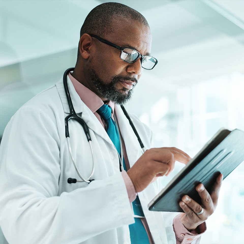 A doctor wearing a white coat and stethoscope uses a tablet device in a bright, modern medical facility.