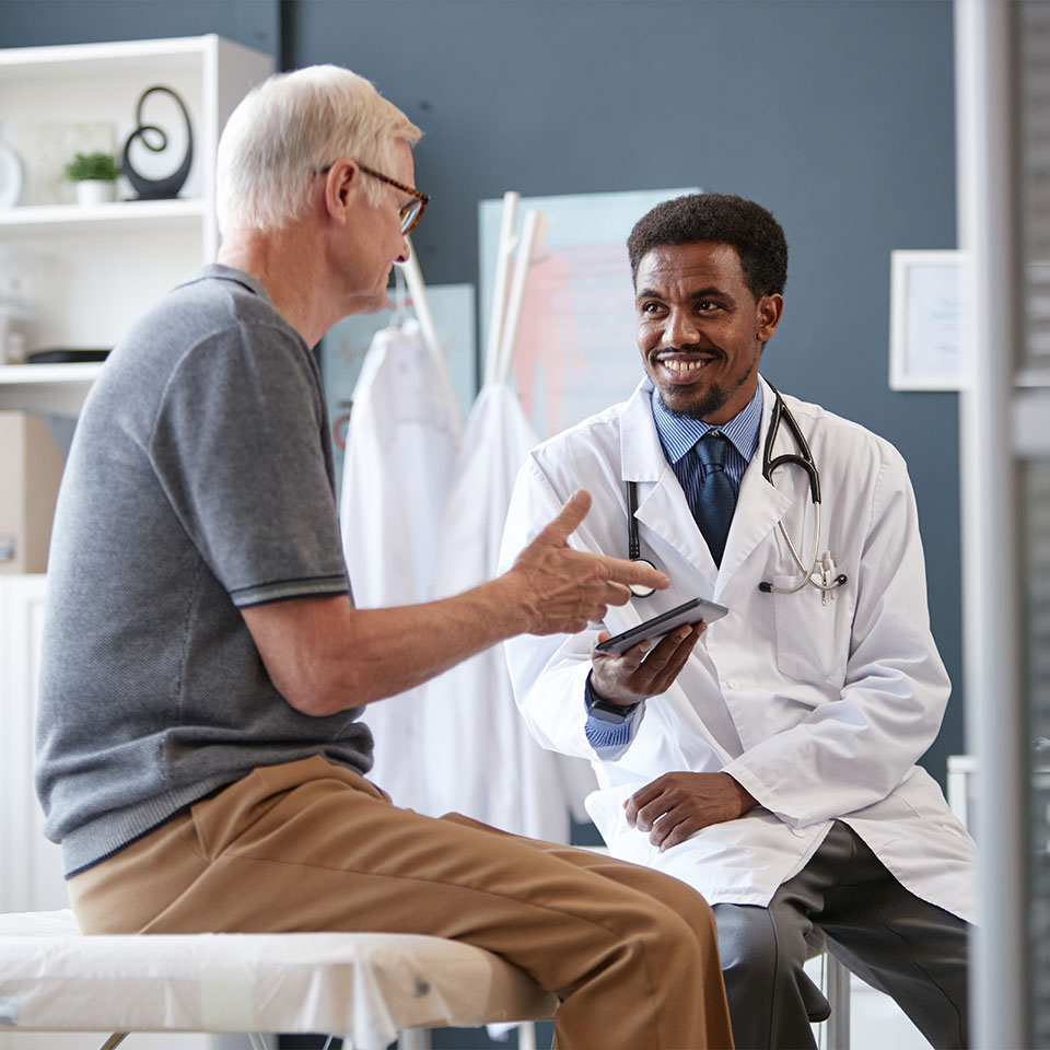 A doctor in a white coat consults with an older male patient, both seated and engaged in conversation in a medical office.