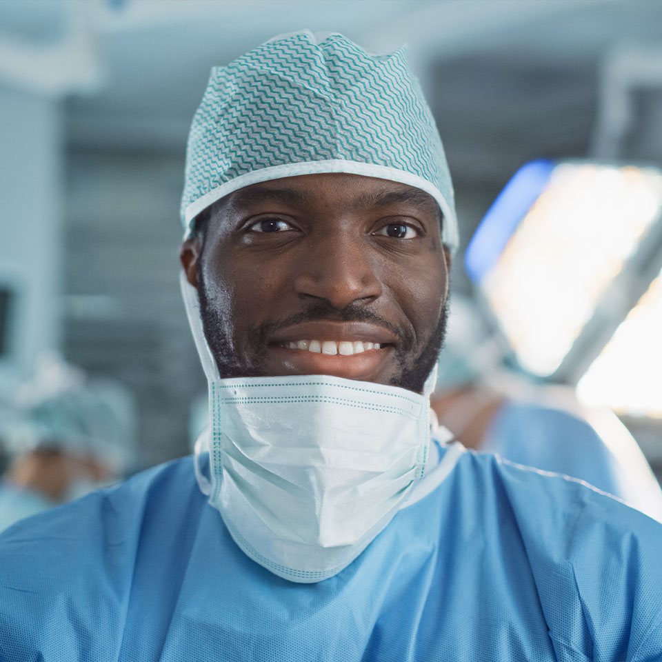 A surgeon wearing scrubs, a surgical cap, and a face mask stands in an operating room, looking at the camera and smiling.