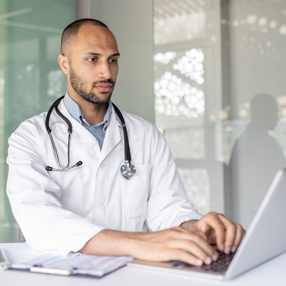 A doctor wearing a white coat and stethoscope types on a laptop at a desk with a notebook and documents nearby.