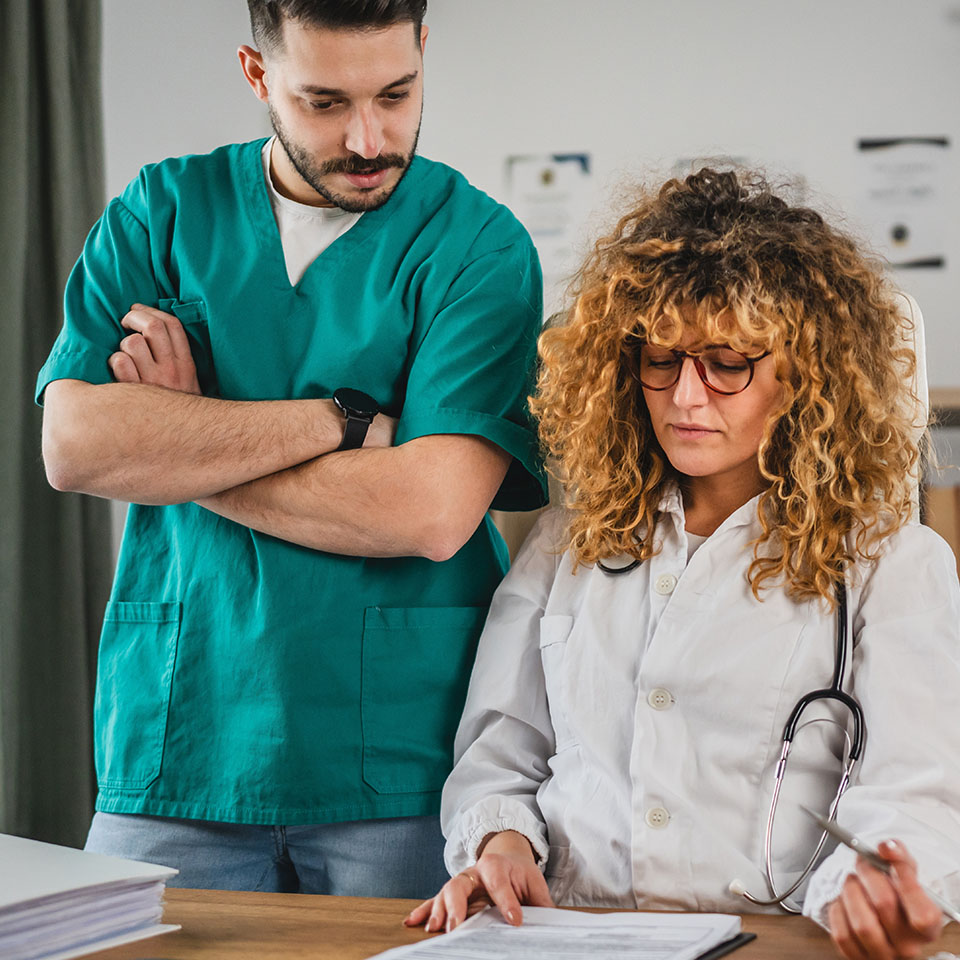 A male healthcare worker in green scrubs and a female doctor in a white coat review documents together at a desk.