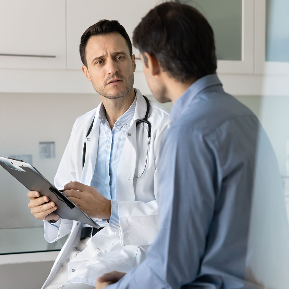 A doctor in a white coat holds a clipboard and speaks with a patient in a blue shirt during a medical consultation in an office.