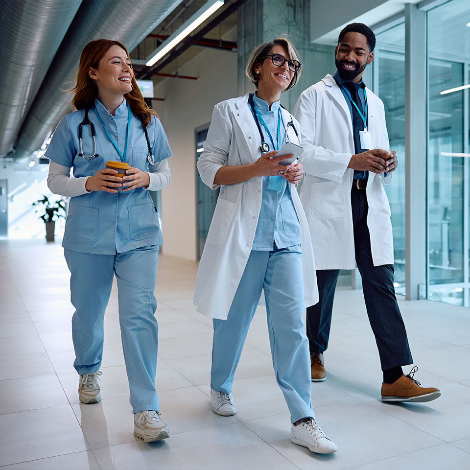 Three healthcare professionals in scrubs and lab coats walk together in a hospital corridor, conversing and smiling.