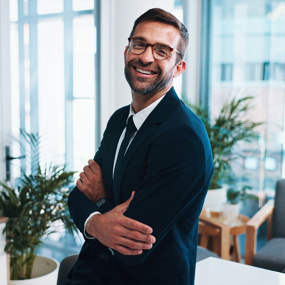 A man in a suit and glasses stands indoors, smiling with arms crossed. Office furniture and plants are visible in the background.
