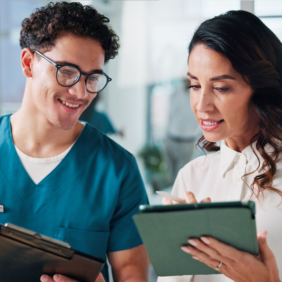 Two professionals, one in medical scrubs and one in business attire, review information together on tablets in a well-lit office setting.
