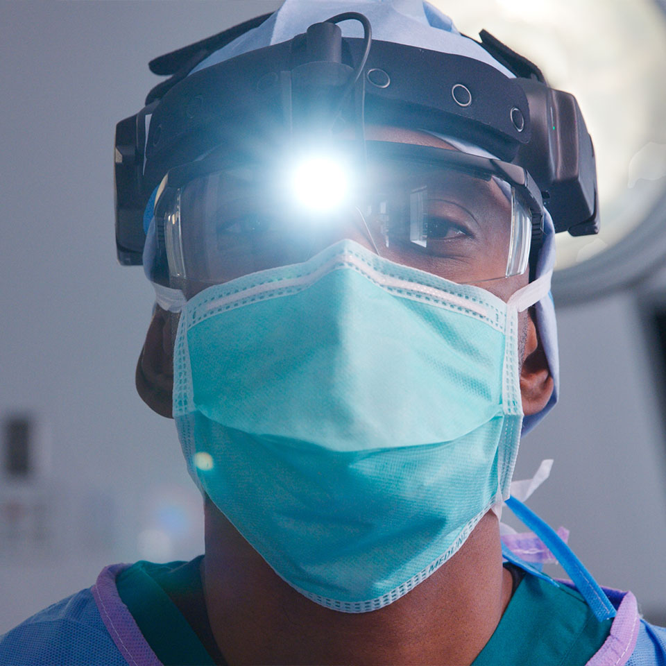 A surgeon wearing surgical mask, protective eyewear, and headlamp looks forward in a brightly lit operating room.