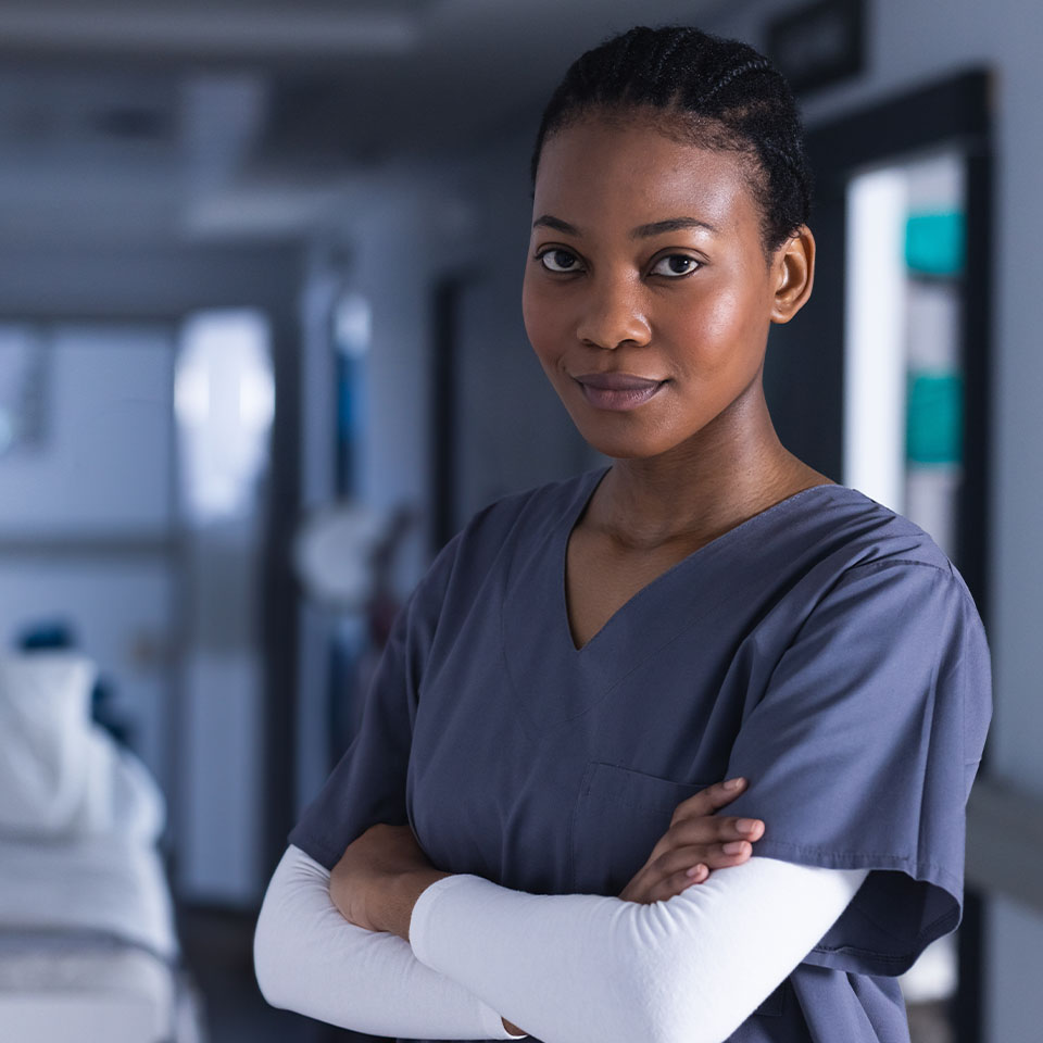 A healthcare worker in gray scrubs stands with folded arms in a hospital hallway, looking at the camera.
