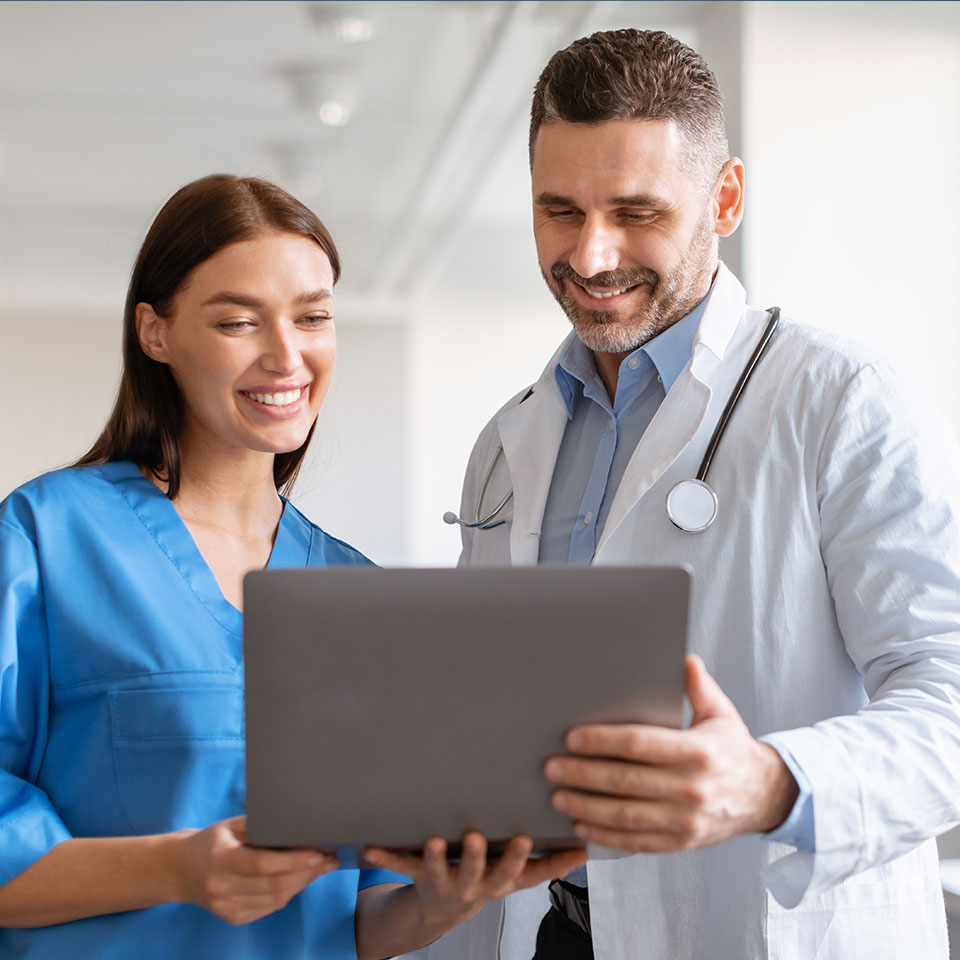 A female nurse and a male doctor stand together, smiling as they look at a laptop screen in a medical setting.