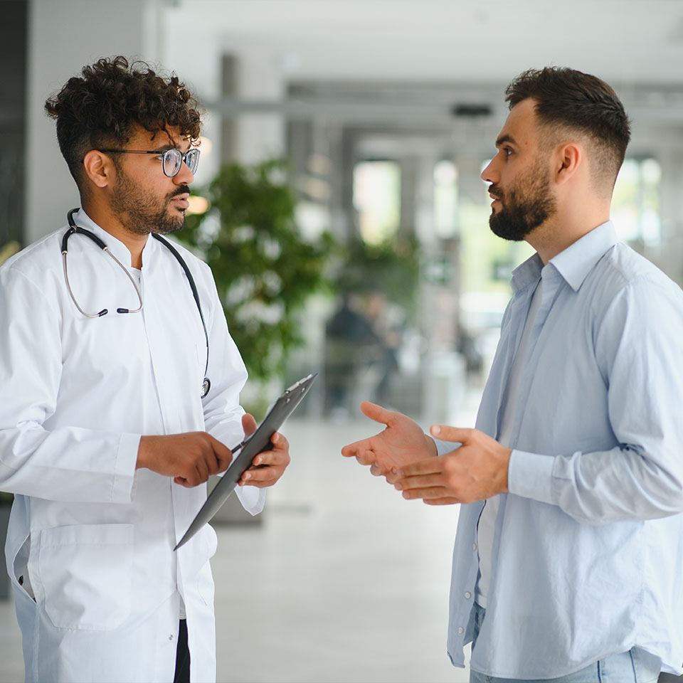 A doctor holding a clipboard listens to a man in a light blue shirt as they have a conversation in a bright, modern indoor setting.