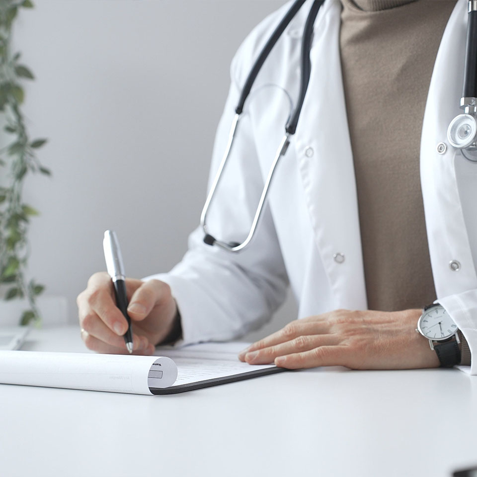 A person wearing a white lab coat and stethoscope writes on a clipboard at a desk.
