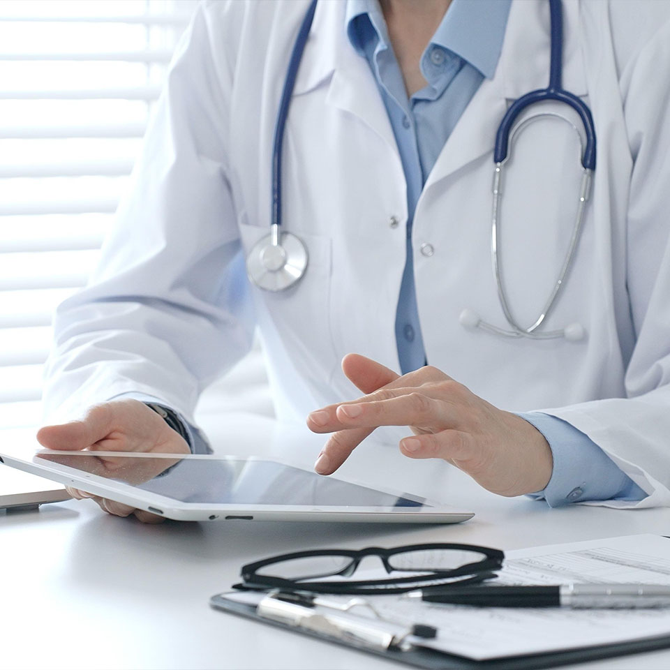 A doctor wearing a white coat and stethoscope uses a tablet at a desk, with eyeglasses and paperwork nearby.