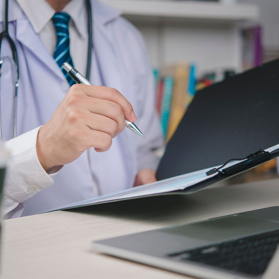 A doctor in a white coat reviews and writes notes on a clipboard at a desk with a laptop.