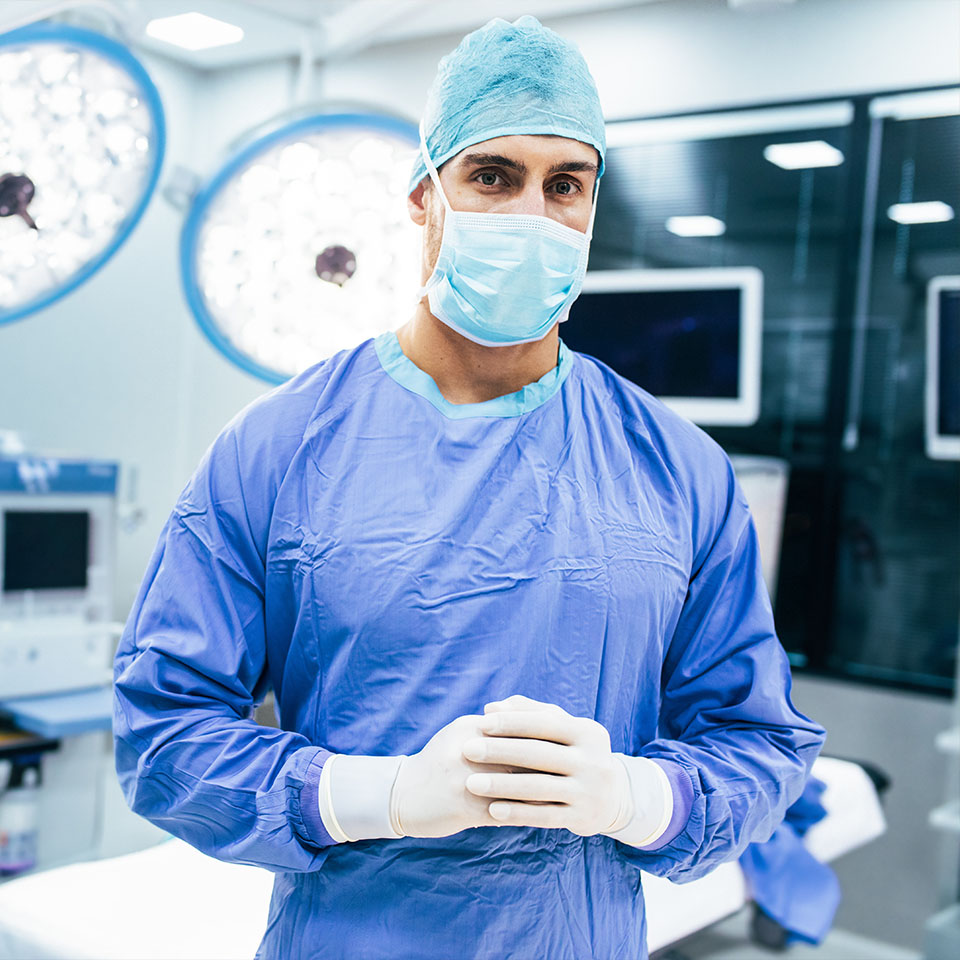 A surgeon wearing a blue gown, cap, mask, and gloves stands in a brightly lit operating room with medical equipment in the background.