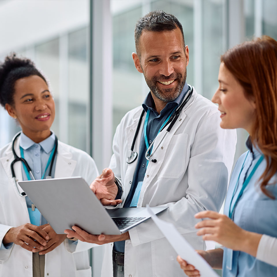 Three doctors in white coats stand together indoors, discussing work while one holds a laptop and another holds papers.