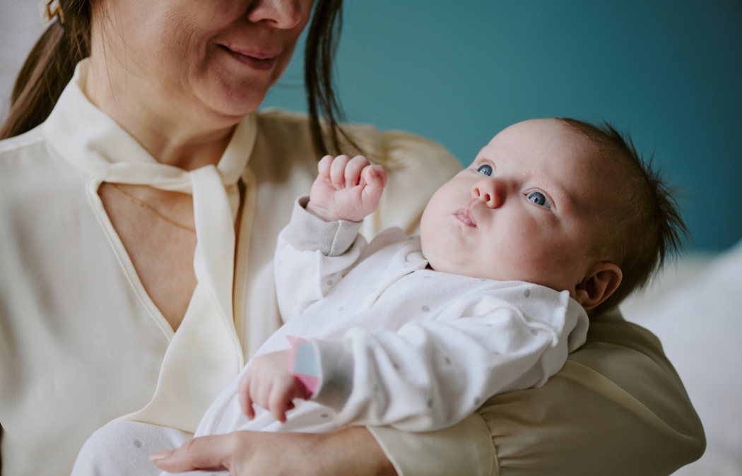 Woman holding newborn baby in vibrant beige room. Baby wearing white onesie, gazing upwards while mother lovingly holding infant close